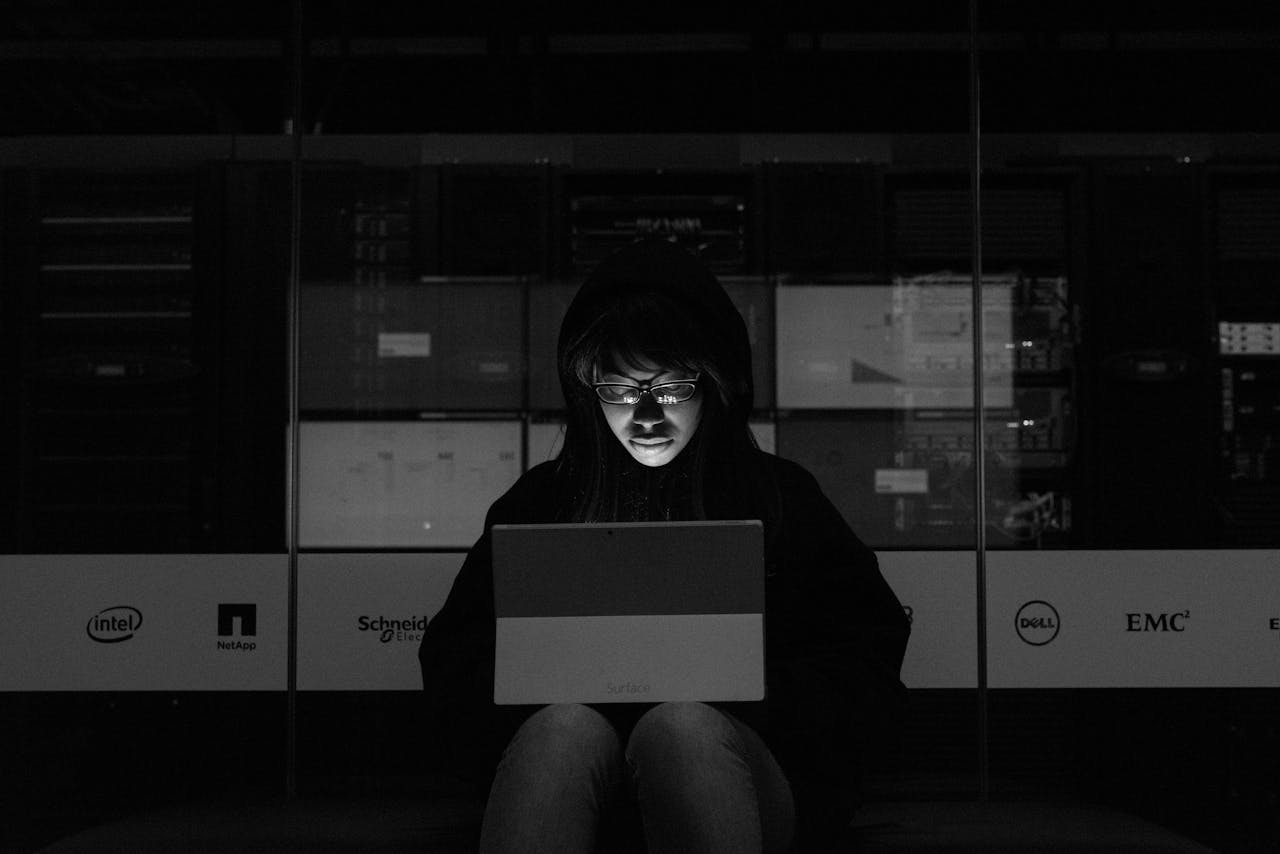 A woman in a dark room uses a laptop, illuminated by its screen, with server equipment in the background.