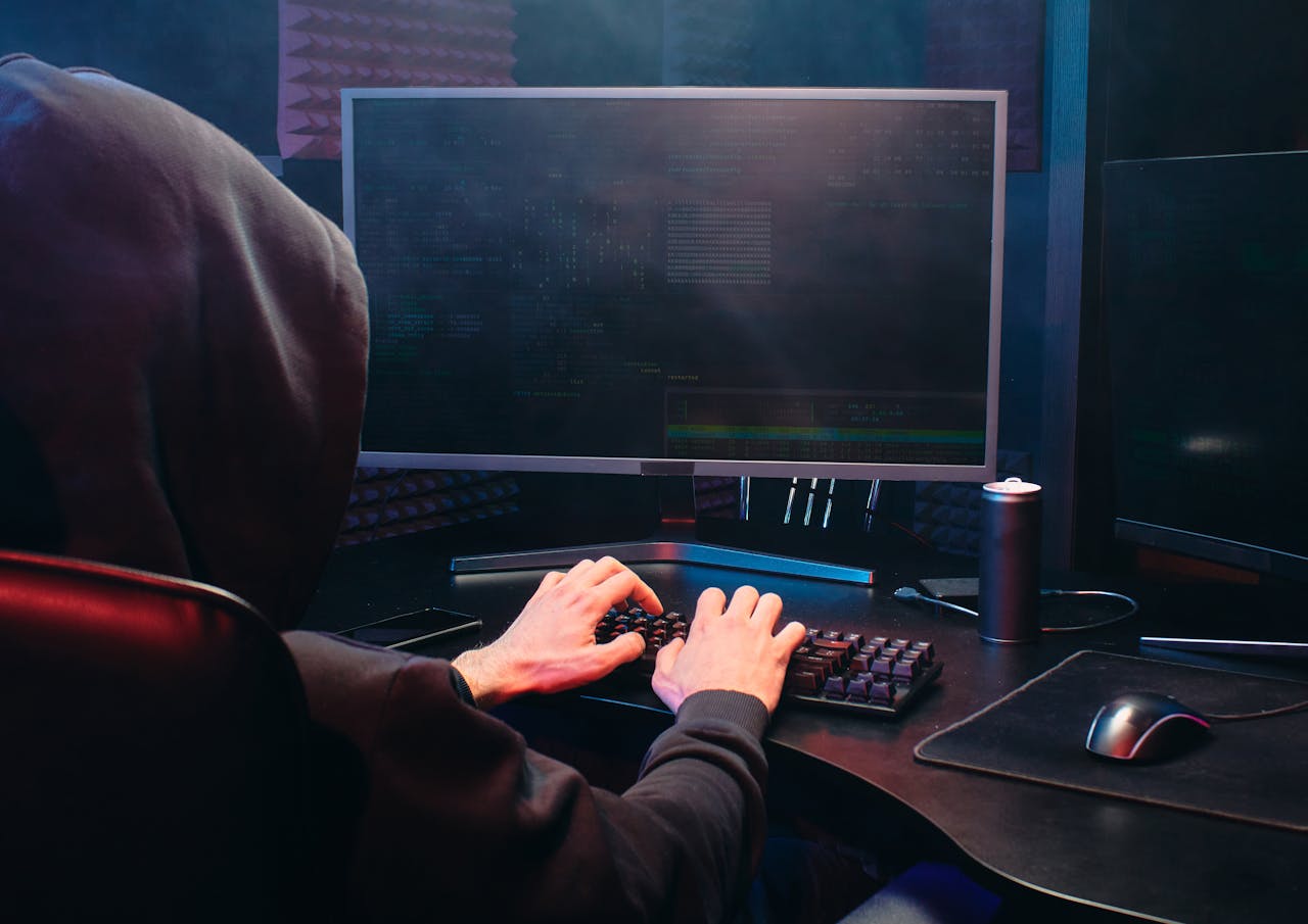 A person in a hoodie sits at a computer screen, engaged in coding or hacking activities.